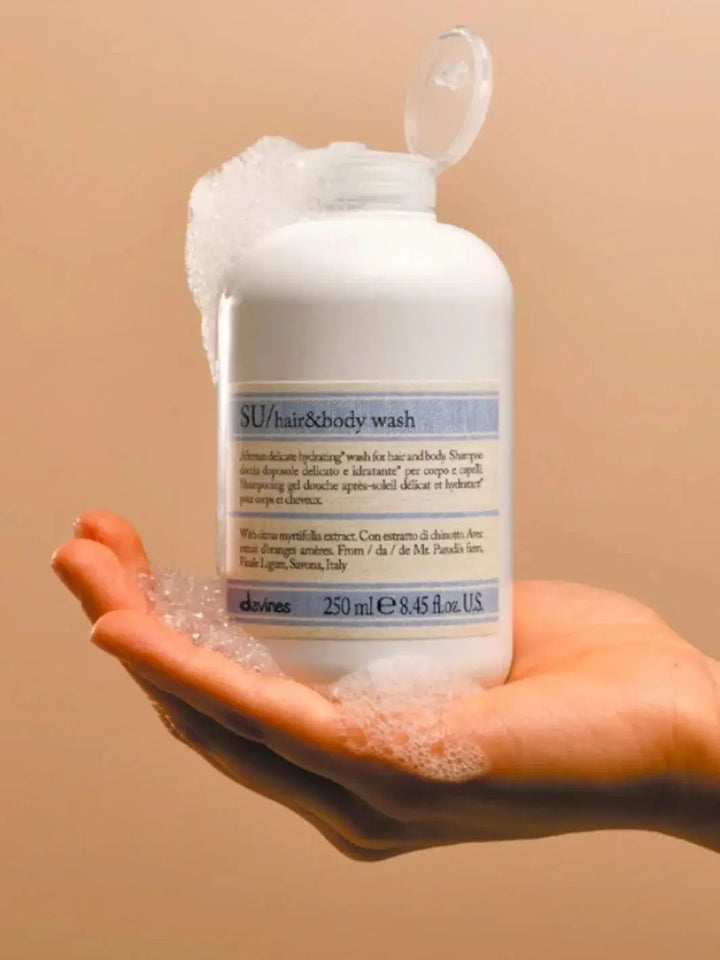 A hand cradles an open bottle of Davines SU Hair and Body Wash covered in frothy soap bubbles against a warm beige background | Twentyseven Toronto
