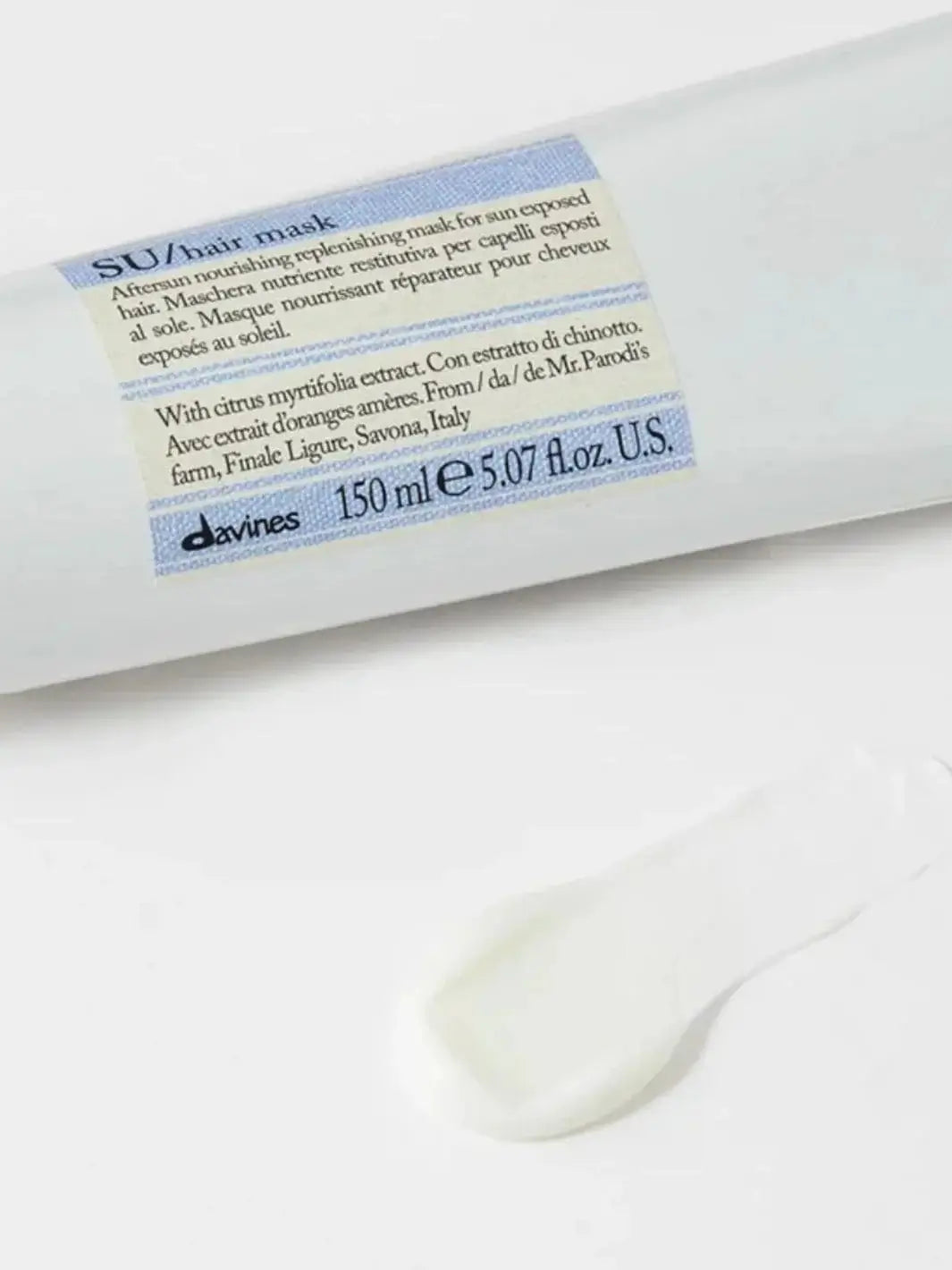 A white squeeze tube of Davines SU Mask After Sun Replenishing Hair Mask lies horizontally on a clean surface next to a smooth smear of the white cream product | Twentyseven Toronto