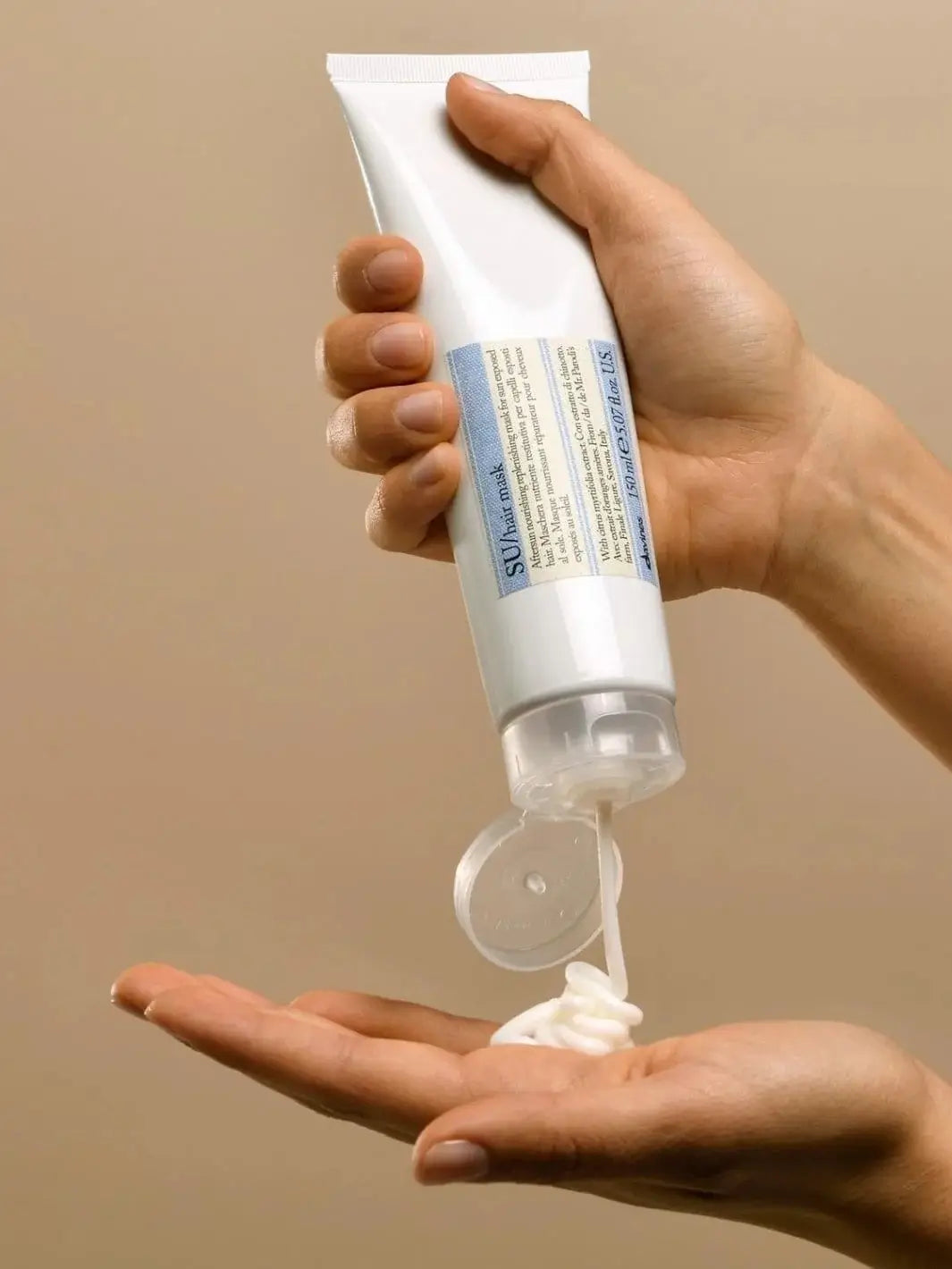 A white tube of Davines SU Mask After Sun Replenishing Hair Mask is held upside down as  white cream is squeezed out onto an open palm | Twentyseven Toronto