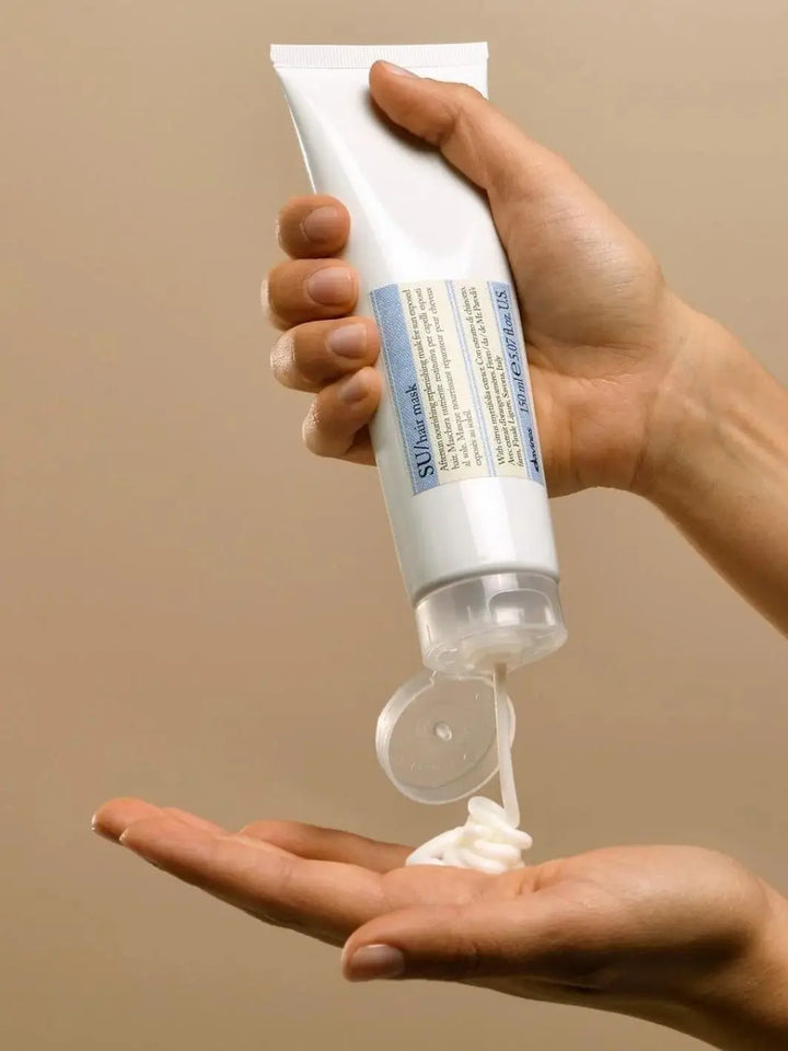 A white tube of Davines SU Mask After Sun Replenishing Hair Mask is held upside down as  white cream is squeezed out onto an open palm | Twentyseven Toronto