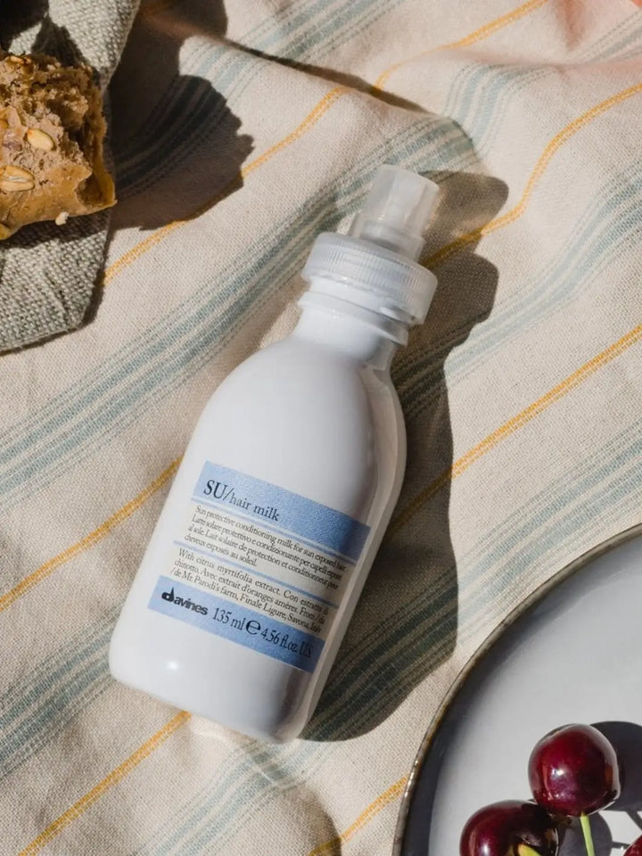 A white spray bottle of Davines SU Milk rests on a striped picnic blanket next to a piece of bread and a plate of cherries | Twentyseven Toronto