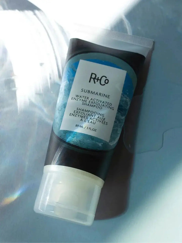 A tube of R+Co SUBMARINE Water Activated Enzyme Exfoliation Shampoo rests on a blue surface reflecting wavy, water-like light patterns | Twentyseven Toronto