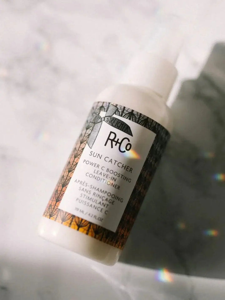A bottle of R+Co SUN CATCHER Leave in Conditioner catching a faint, soft rainbow prism of light across its label | Twentyseven Toronto