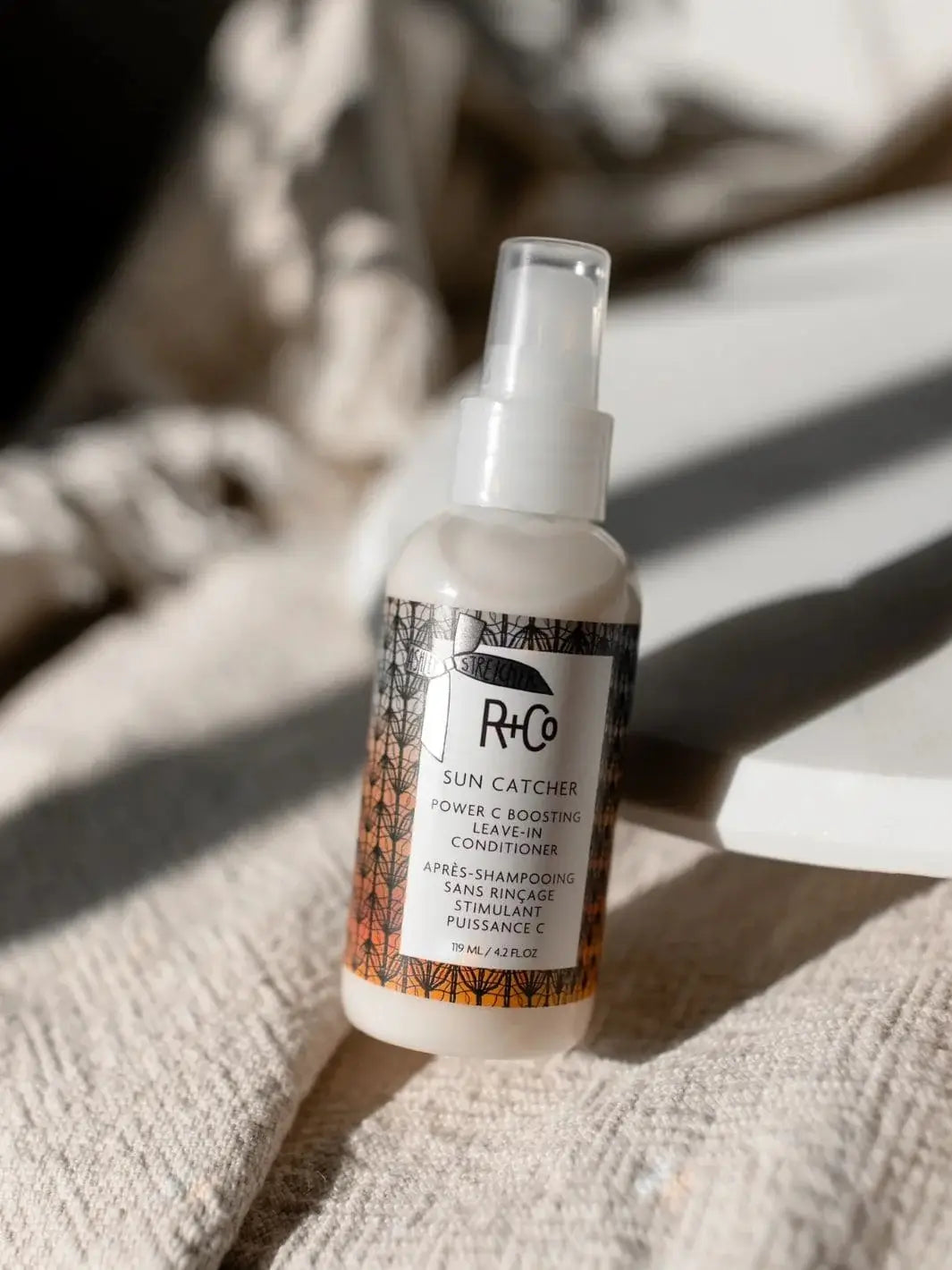 A bottle of R+Co SUN CATCHER Leave in Conditioner stands upright on a textured cream blanket under bright, casting shadows | Twentyseven Toronto