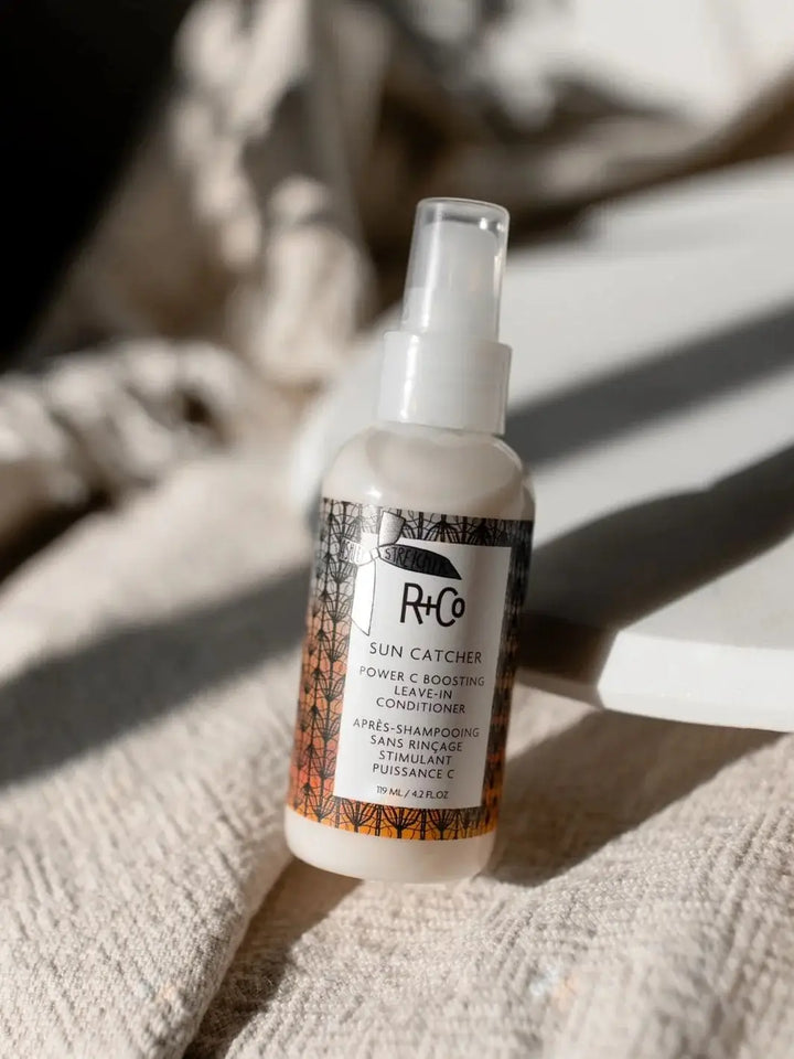 A bottle of R+Co SUN CATCHER Leave in Conditioner stands upright on a textured cream blanket under bright, casting shadows | Twentyseven Toronto