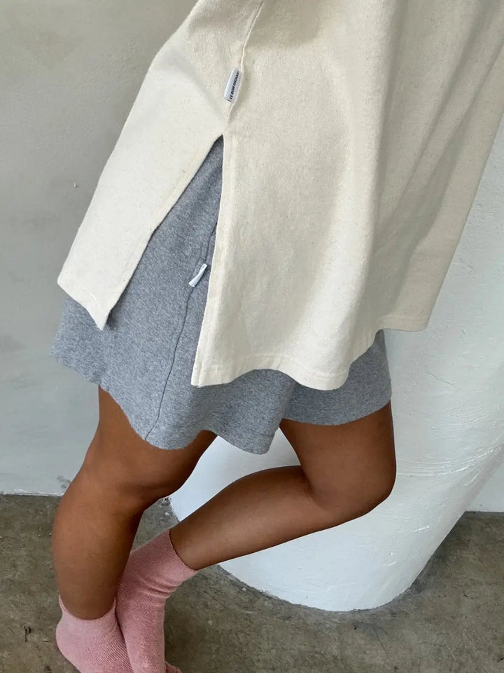 Person wearing a beige 'Sunday Tee' sweatshirt in 'Naturel' over gray shorts with pink socks from Le Bon Shoppe, standing against a neutral background | Twentyseven Toronto