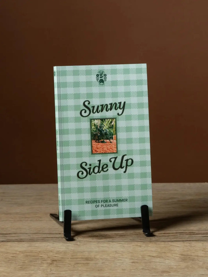 The green gingham front cover of the Flamingo Estate Sunny Side Up Recipe Book resting on a small black display stand on a wooden surface | Twentyseven