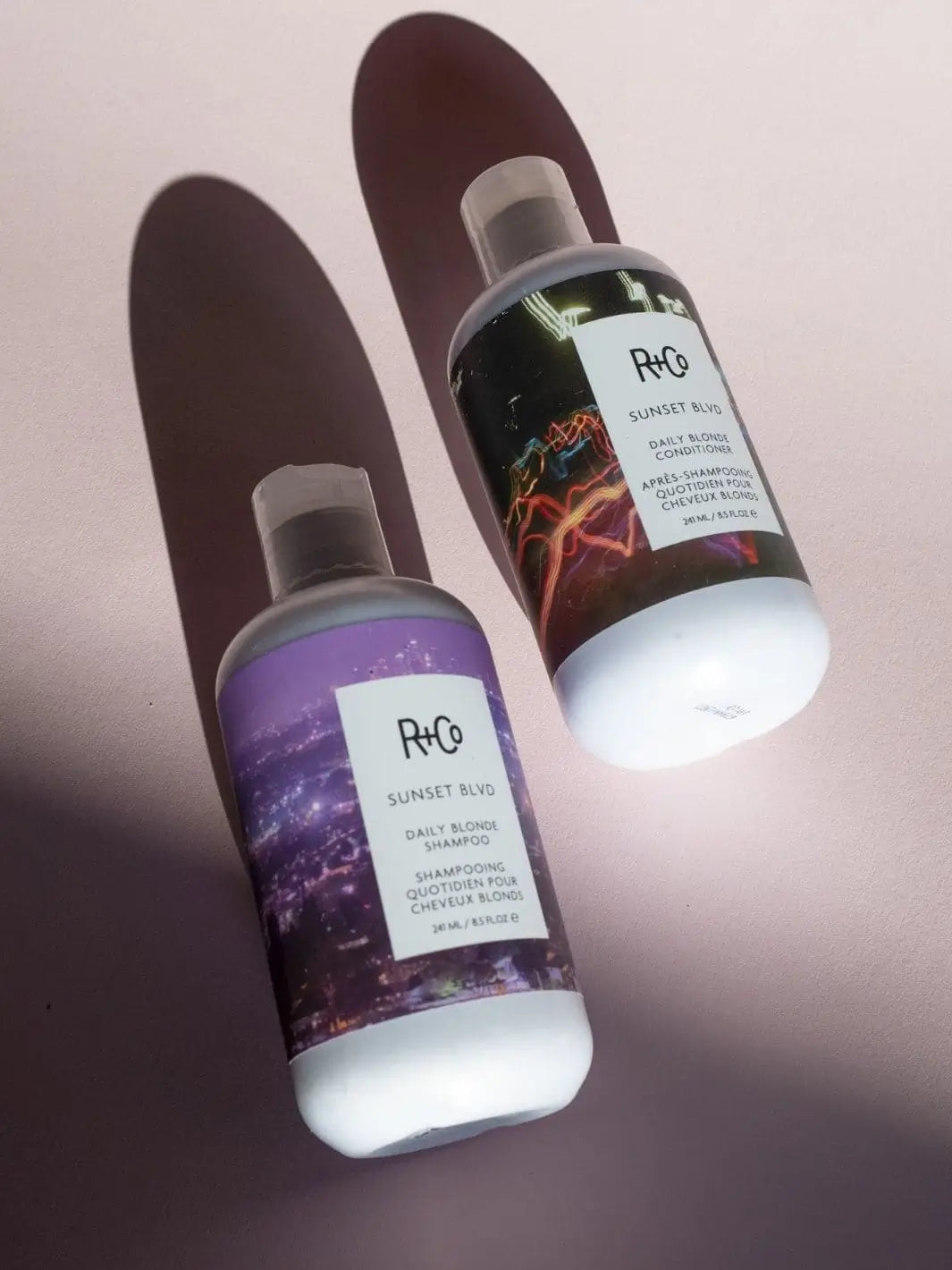 A bottle of R+Co SUNSET BLVD Blonde Shampoo is shown next to its matching SUNSET BLVD Blonde Conditioner on a dusty pink background | Twentyseven Toronto