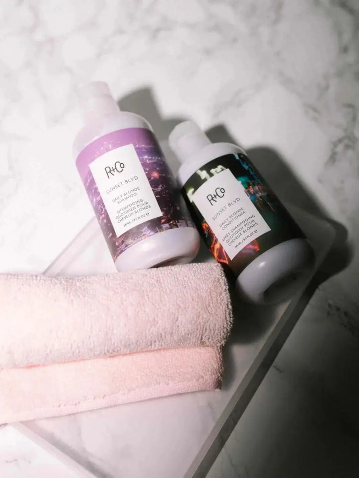 A bottle of R+Co SUNSET BLVD Blonde Shampoo rests beside R+Co SUNSET BLVD Conditioner and a folded pink washcloth on a marble counter | Twentyseven Toronto