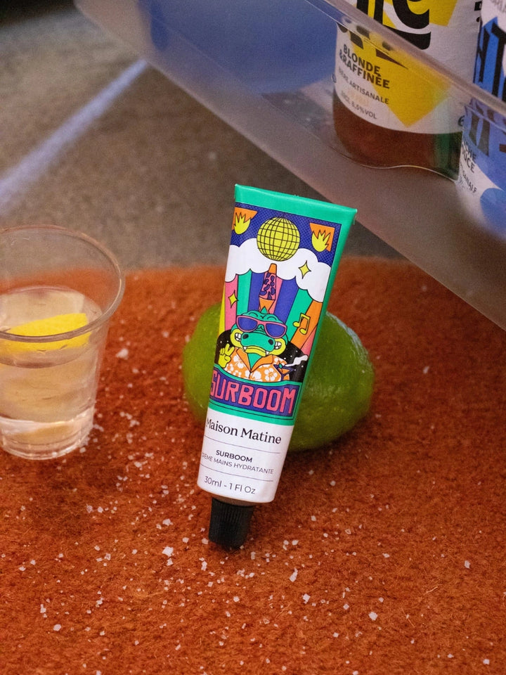 Maison Matine Surboom Hand Cream | Twentyseven Toronto | A tube of Maison Matine Surboom Hand Cream on a textured surface, with a glass, lime and bottle in the background.