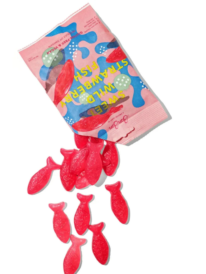 Bright red, fish-shaped gummy candies spill out from an open pink and blue pouch of Sweet Wild Strawberry Fish from BonBon NYC onto a clean white surface | Twentyseven Toronto