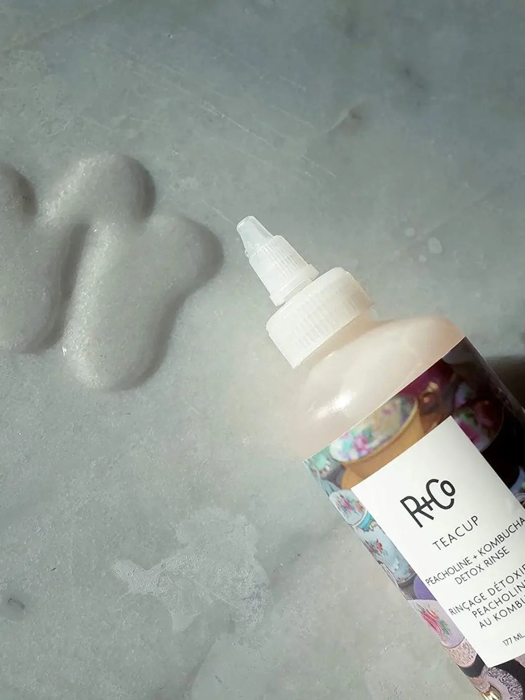 A close-up shot shows a bottle of R+Co TEACUP Peacholine + Kombucha Detox Rinse lying on a light grey marbled surface next to a puddle of the liquid product | Twentyseven Toronto