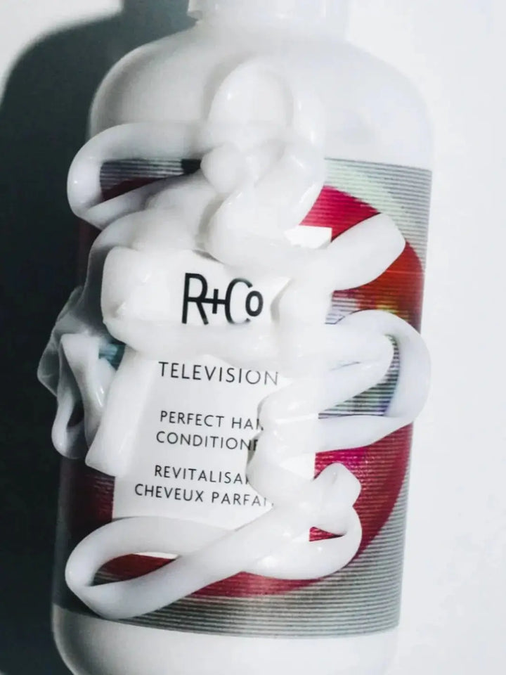 A thick ribbon of white creamy product is swirled directly over the front label of a bottle of R+Co TELEVISION Perfect Hair Conditioner | Twentyseven Toronto