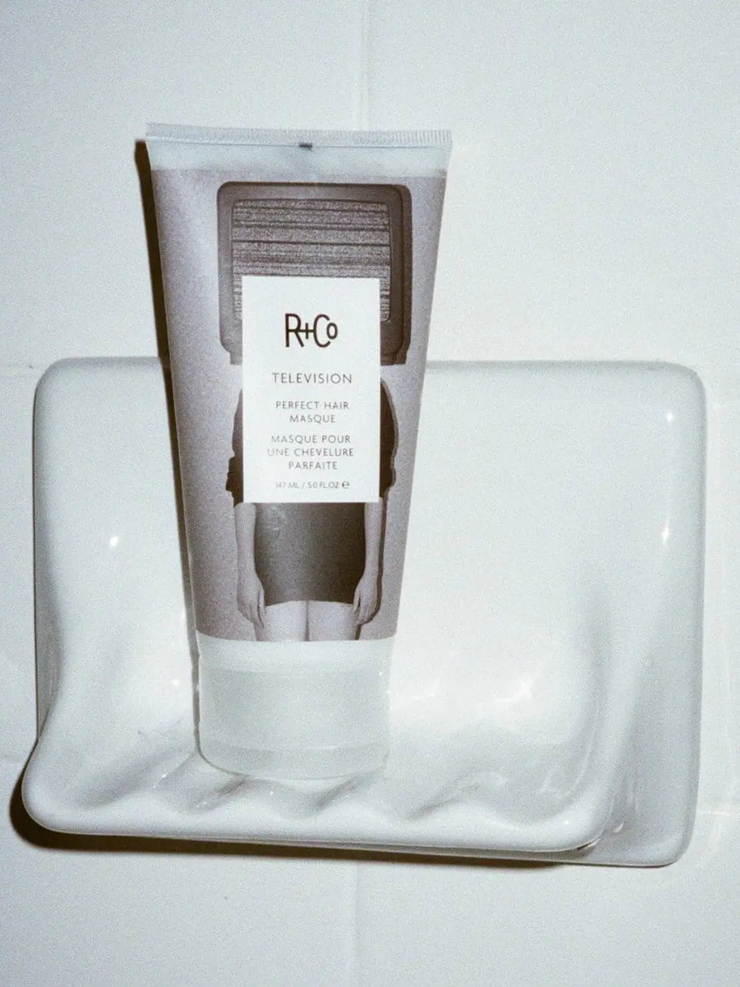 A squeeze tube of R+Co TELEVISION Perfect Hair Masque stands upright on a white ceramic shower soap dish | Twentyseven Toronto