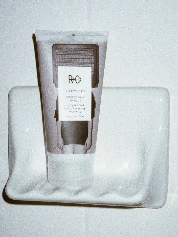 A squeeze tube of R+Co TELEVISION Perfect Hair Masque stands upright on a white ceramic shower soap dish | Twentyseven Toronto