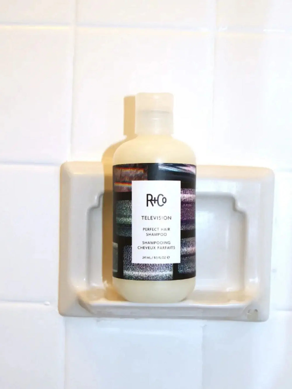 A bottle of R+Co TELEVISION Perfect Hair Shampoo sits upright in a built-in ceramic shower shelf | Twentyseven Toronto