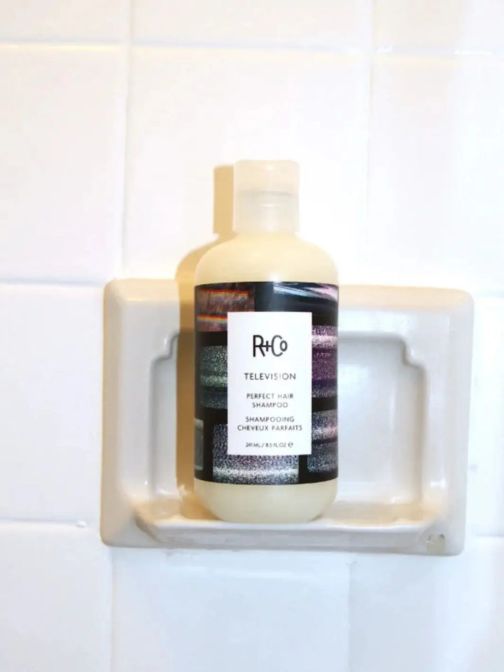 A bottle of R+Co TELEVISION Perfect Hair Shampoo sits upright in a built-in ceramic shower shelf | Twentyseven Toronto
