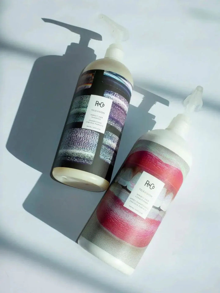 Two large pump bottles of R+Co TELEVISION Perfect Hair Shampoo and Conditioner Liter Size resting side-by-side on a light surface casting soft shadows | Twentyseven Toronto