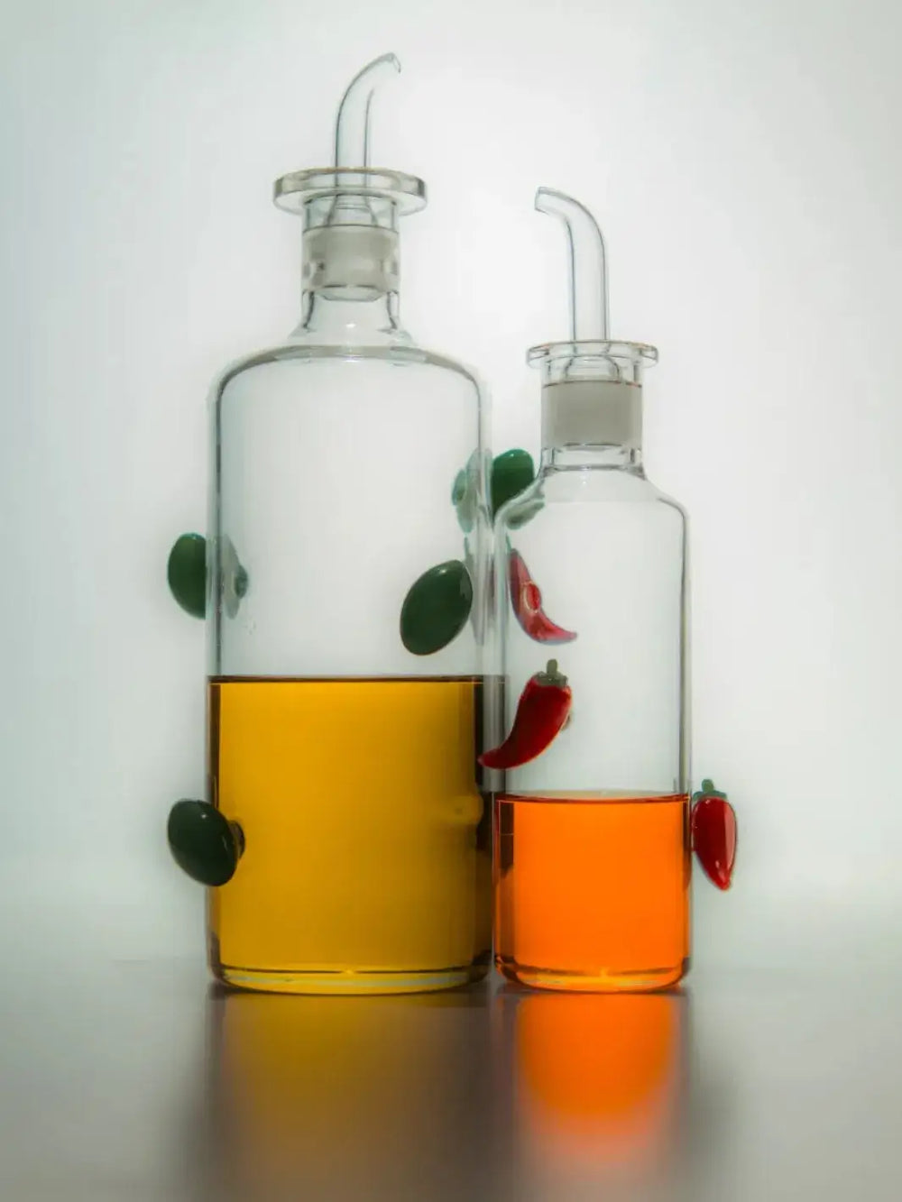 Two clear glass bottles from Maison Balzac with liquid, one olive oil bottle containing a olive oil and the chilli bottle containing chilli oil liquid, on a reflective surface | Twentyseven Toronto