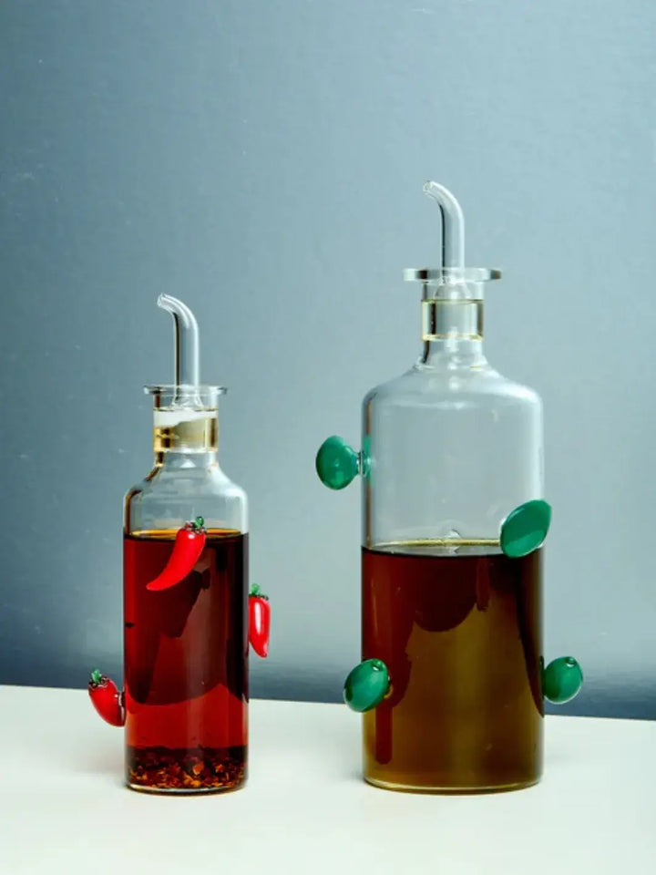 Two clear glass bottles from Maison Balzac with liquid, one olive oil bottle containing a olive oil and the chilli bottle containing chilli oil liquid, on a light blue background | Twentyseven Toronto