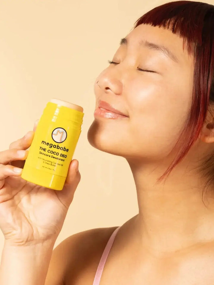 A woman smiling with closed eyes holds a yellow stick of skincare deodorant near her face to emphasize the pleasing scent of the Megababe The Coco Deo | Twentyseven Toronto