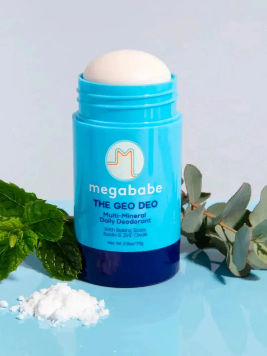 An opened bright blue deodorant stick stands vertically on a light blue surface next to fresh mint leaves, a eucalyptus sprig, and a small pile of white powder to showcase the ingredients of Megababe The Geo Deo | Twentyseven Toronto