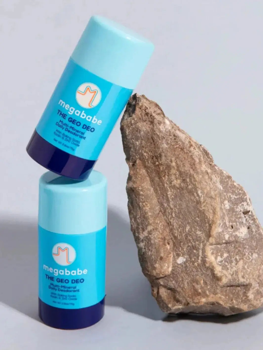 One upright container and one tilted container of Megababe The Geo Deo are arranged symmetrically against a large, rough natural stone on a neutral grey background to emphasize the mineral-based formula | Twentyseven Toronto