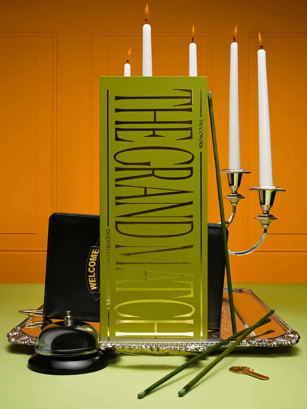 A large, stylishly designed green box of The Grand Match from Piecework Puzzles stands vertically on a silver tray alongside extra-long matchsticks, a candelabra, and a hotel bell | Twentyseven Toronto