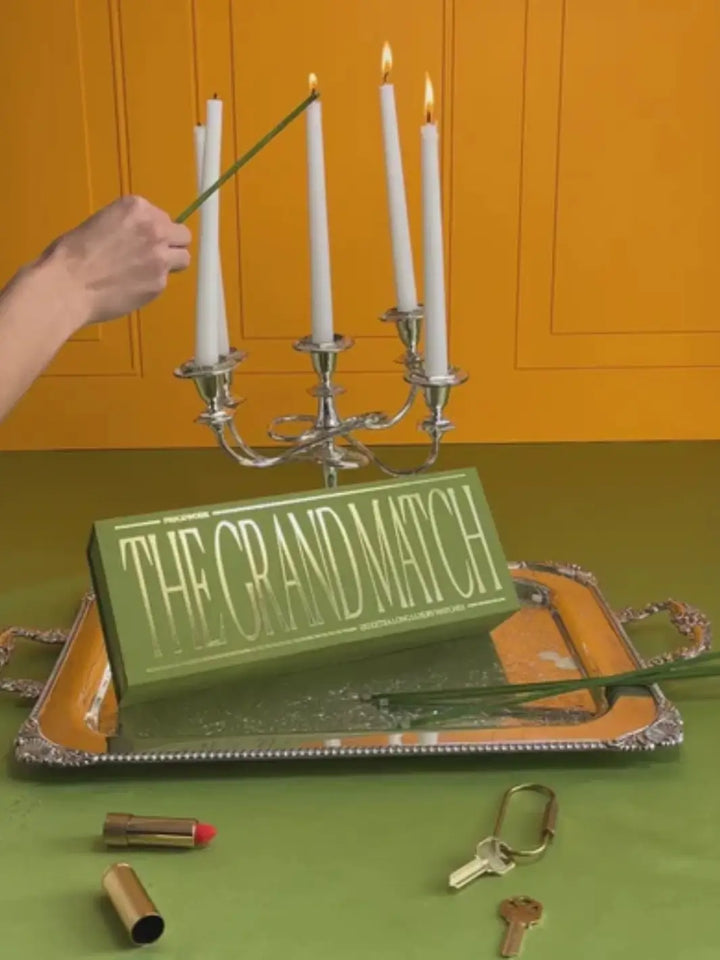 A hand uses an extra-long green matchstick to light white candles in a silver candelabra next to a green box of The Grand Match from Piecework Puzzles | Twentyseven Toronto