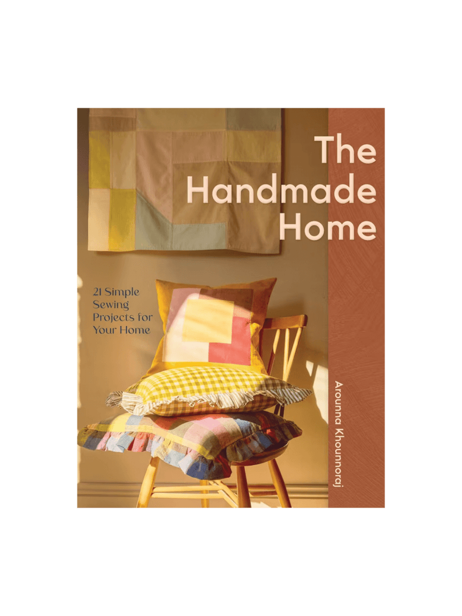 Front cover of the book The Handmade Home by Arounna Khounnoraj, featuring a warm, sunlit interior with a stack of colourful, patterned handmade pillows on a wooden chair | Twentyseven Toronto