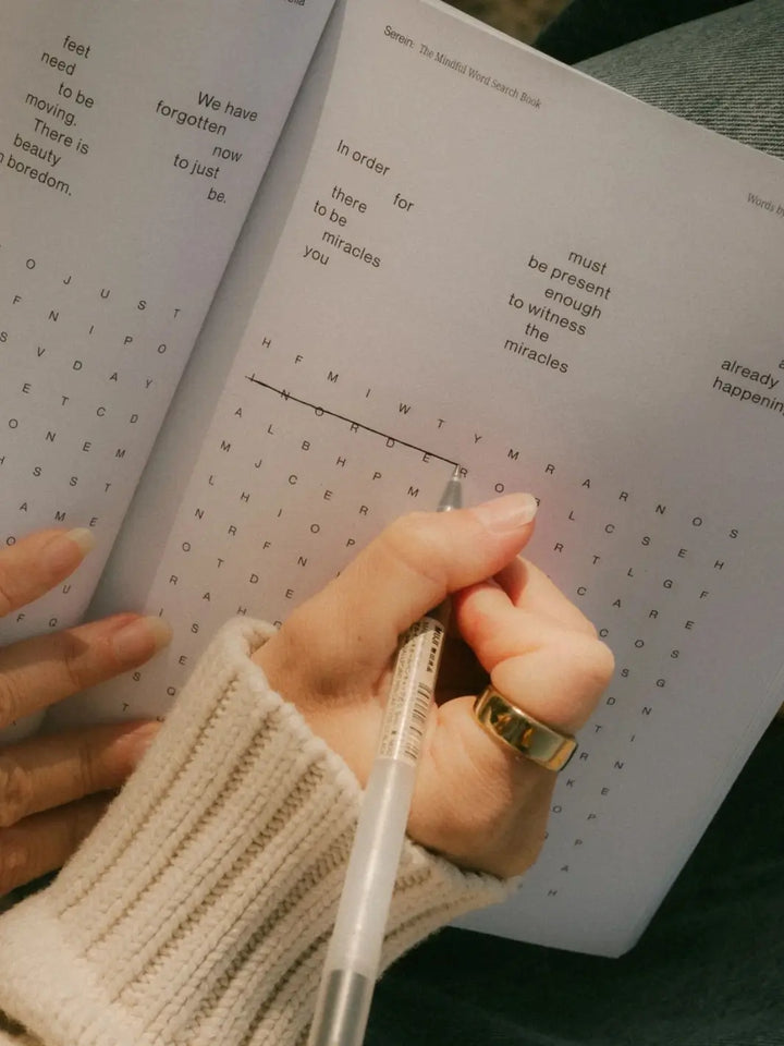 A close-up shot shows a hand wearing a thick gold ring using a pen to solve a puzzle inside an open copy of Serein The Mindful Word Search Book | Twentyseven Toronto