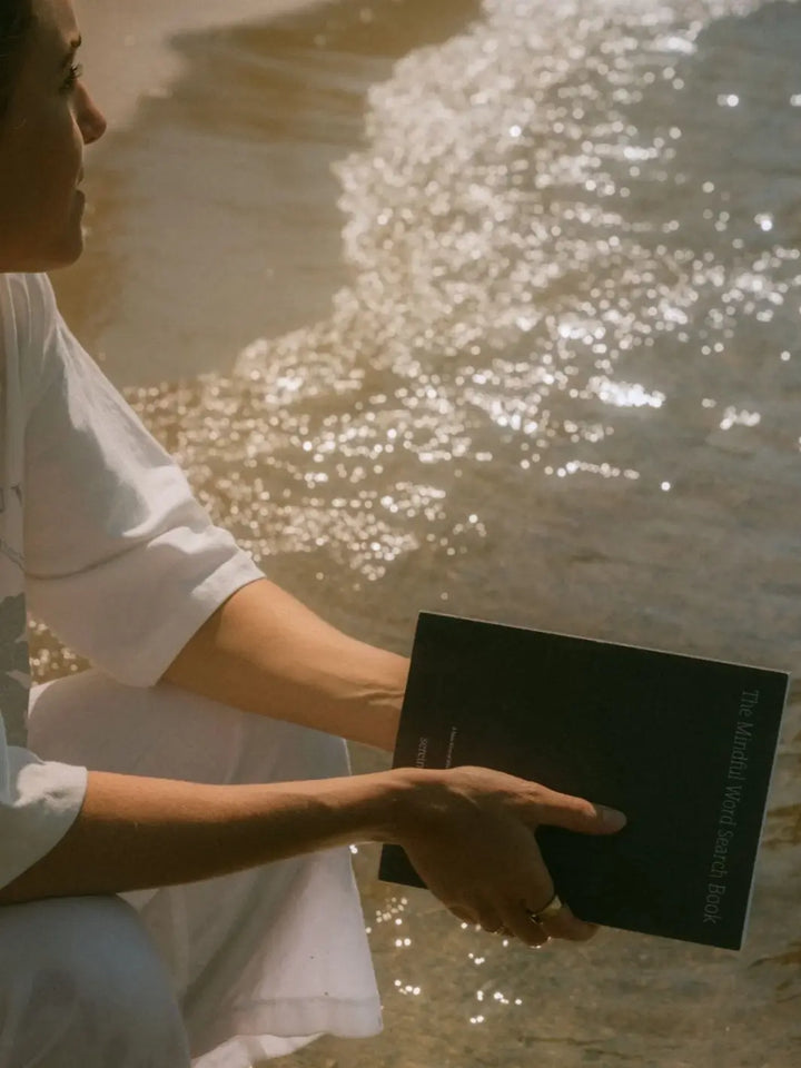 A person sitting by the edge of sparkling water holds a dark-covered copy of Serein The Mindful Word Search Book | Twentyseven Toronto