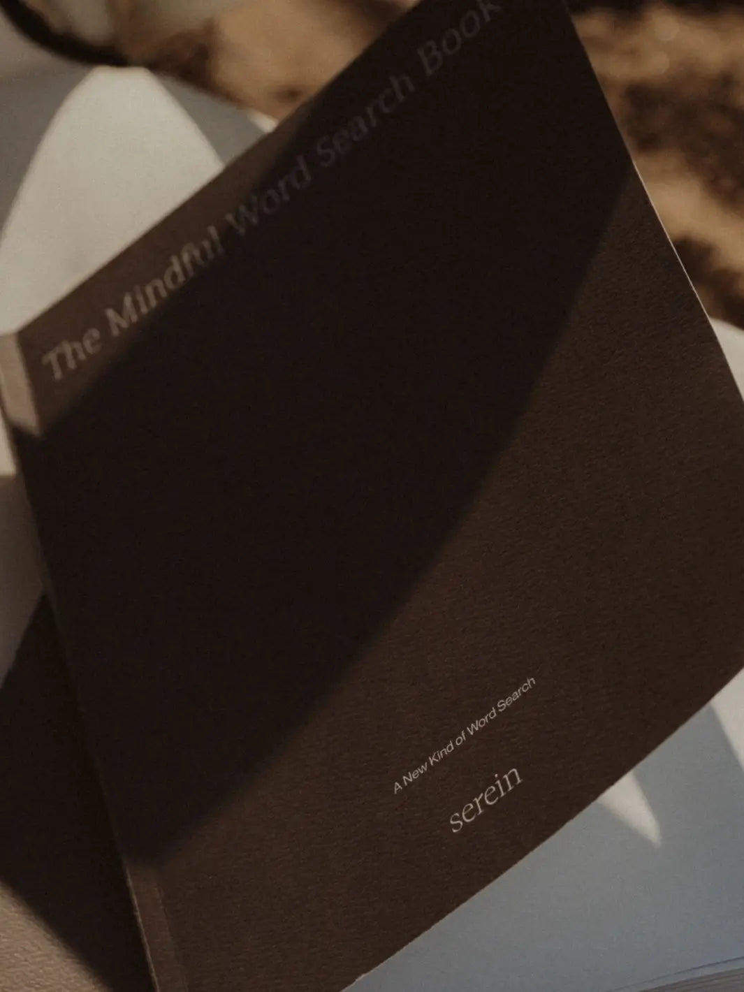 An angled close-up highlights the minimal, textured brown cover of Serein The Mindful Word Search Book casting a soft shadow | Twentyseven Toronto