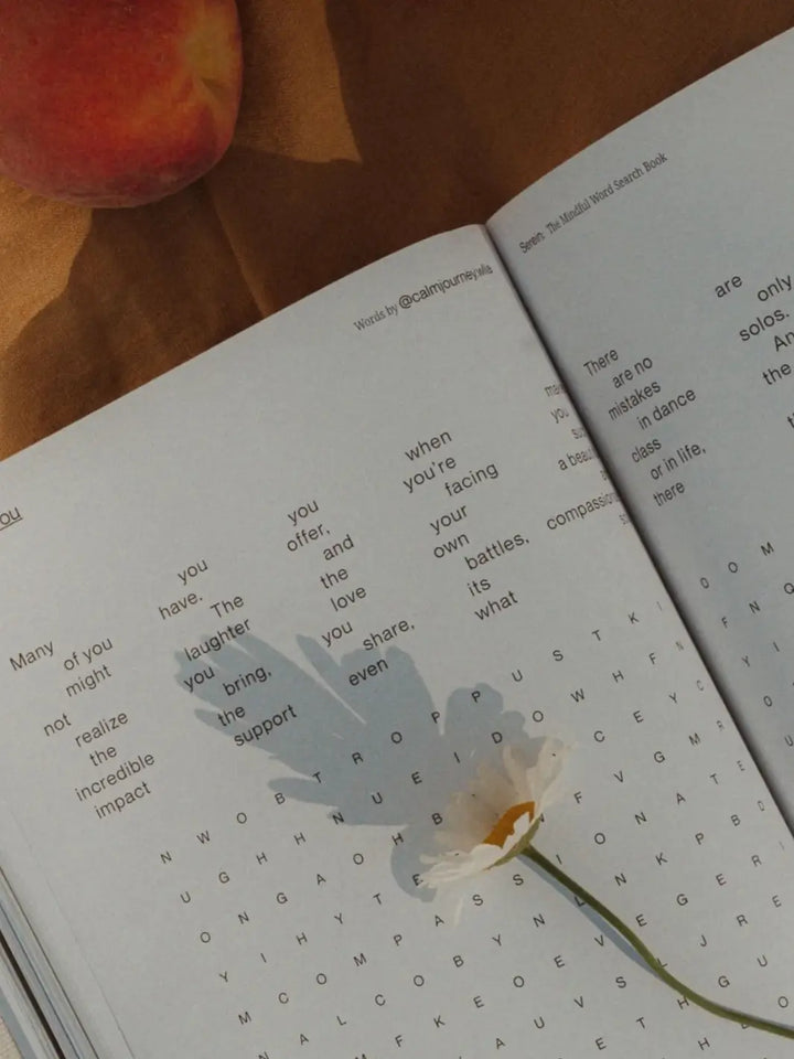 A single white daisy rests across an open page of Serein The Mindful Word Search Book next to a fresh peach | Twentyseven Toronto