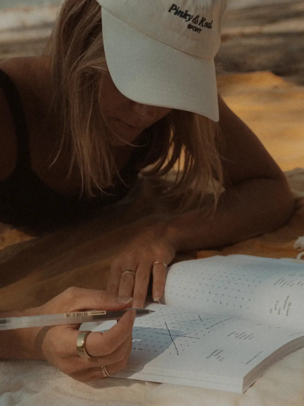 A person wearing a white baseball cap and knit sweater lies down outdoors while filling out a puzzle in Serein The Mindful Word Search Book | Twentyseven Toronto