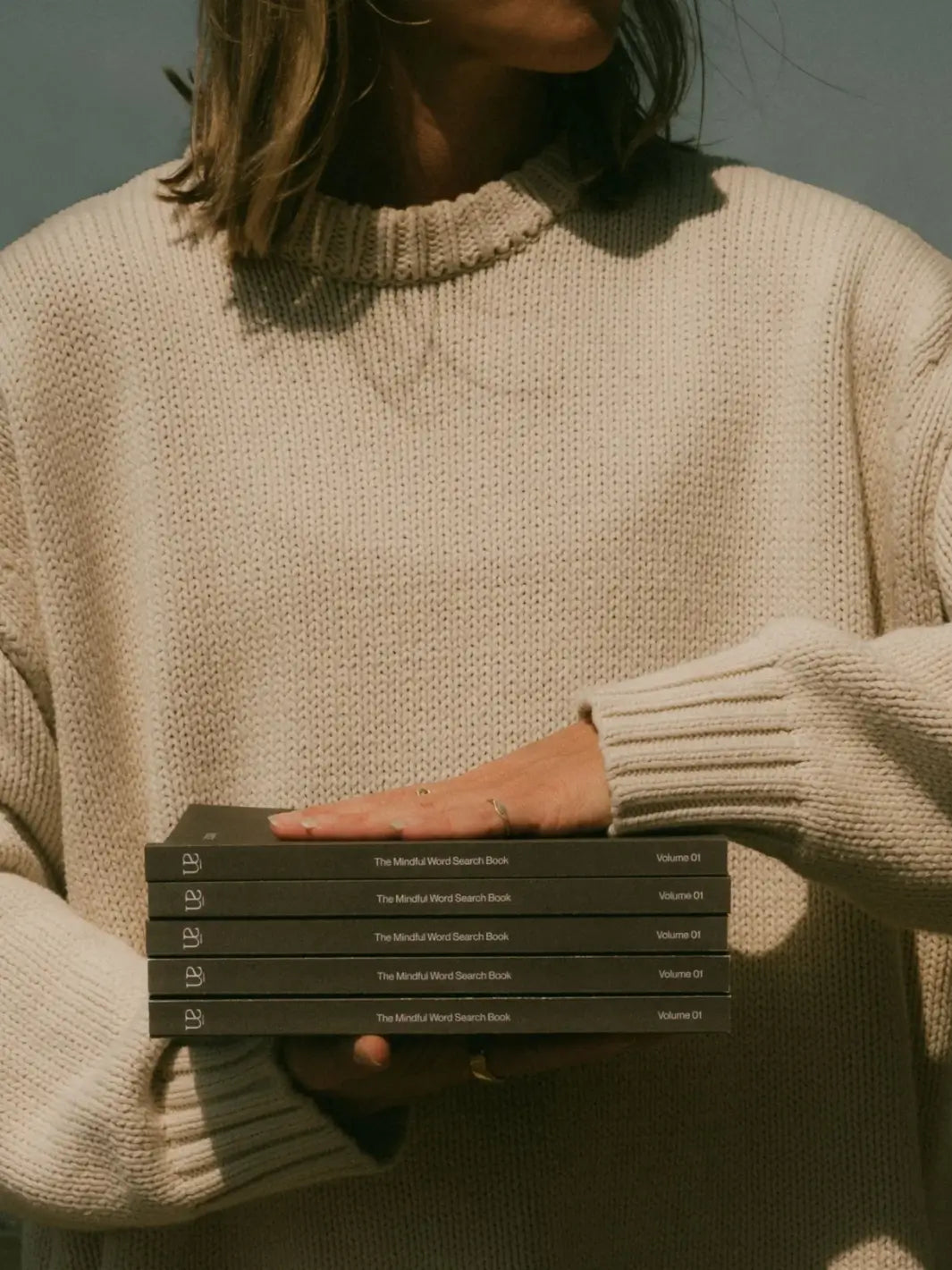 A person dressed in a chunky cream knit sweater holds a stack of five copies of Serein The Mindful Word Search Book Volume 01 | Twentyseven Toronto