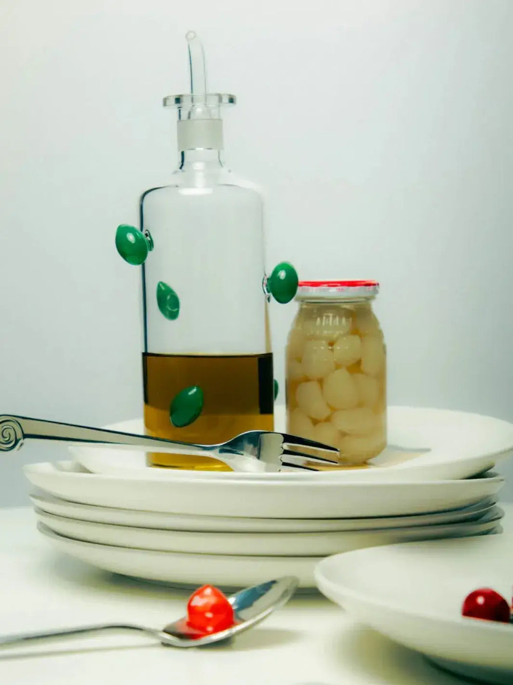 Glass olive oil dispenser bottle from Maison Balzac decorated with green olives filled with oil on top of white ceramic plated | Twentyseven Toronto