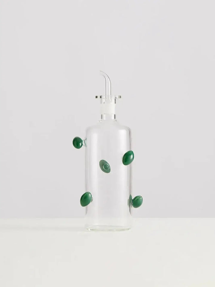 Glass olive dispenser bottle from Maison Balzac decorated with green olives on a white background | Twentyseven Toronto
