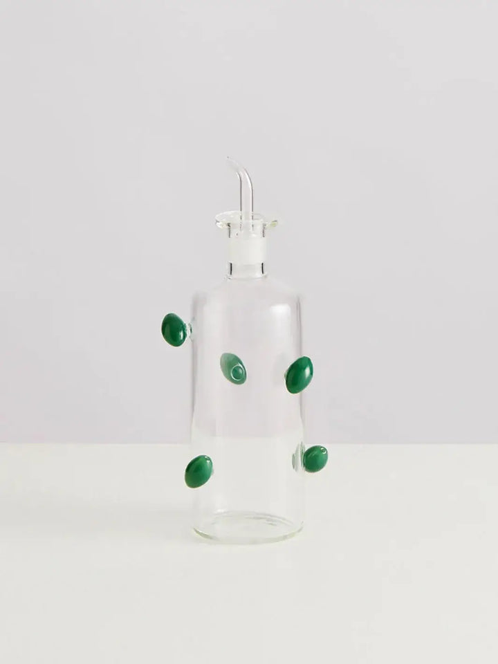 Glass olive dispenser bottle from Maison Balzac decorated with green olives on a white background | Twentyseven Toronto