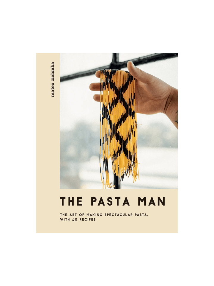 Front cover of The Pasta Man: The Art of Making Spectacular Pasta by Mateo Zielonka features a hand holding up long strands of intricately patterned yellow and black pasta against a bright window | Twentyseven Toronto