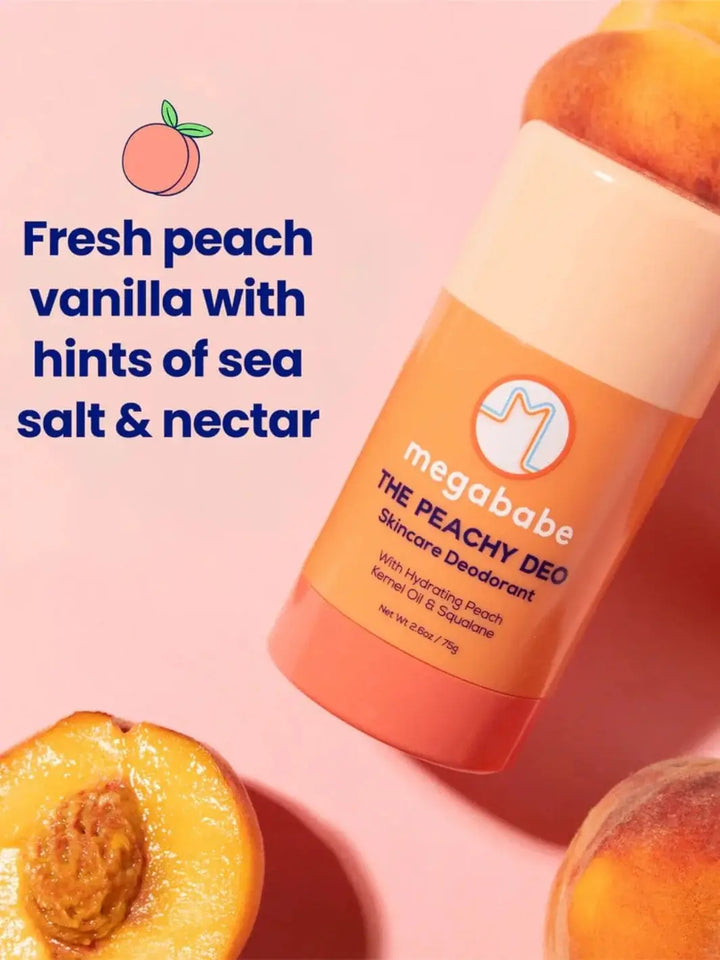An orange stick of Megababe The Peachy Deo is styled flat lay next to fresh whole and halved peaches against a pink background | Twentyseven Toronto