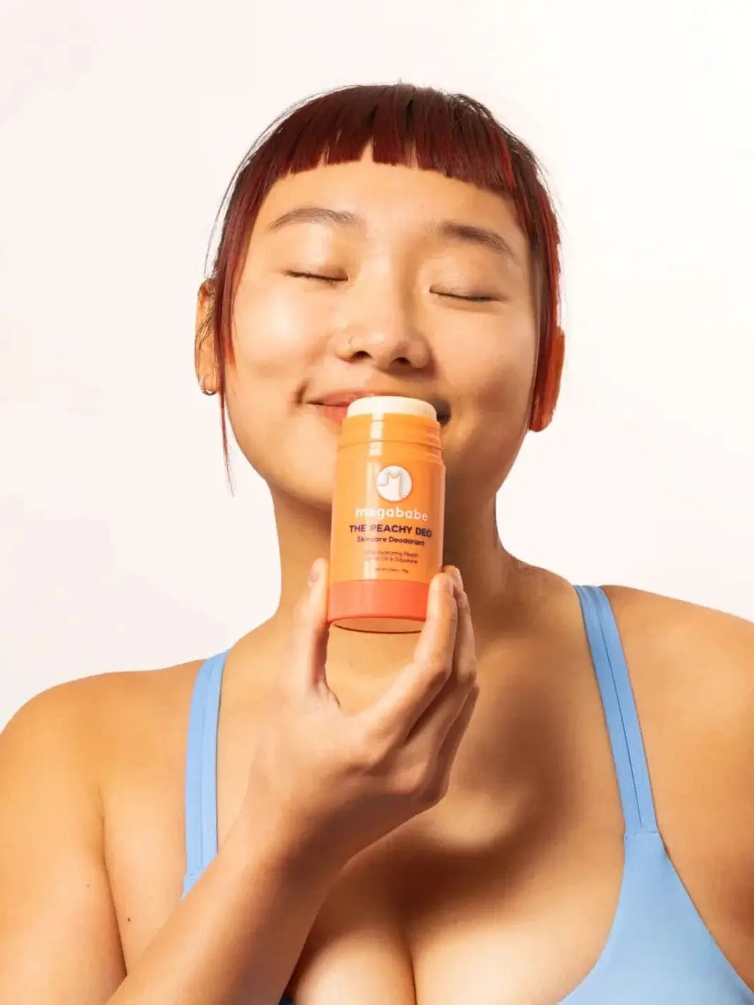 A smiling person with closed eyes holds a stick of Megababe The Peachy Deo up near their face | Twentyseven Toronto