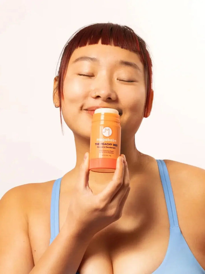 A smiling person with closed eyes holds a stick of Megababe The Peachy Deo up near their face | Twentyseven Toronto