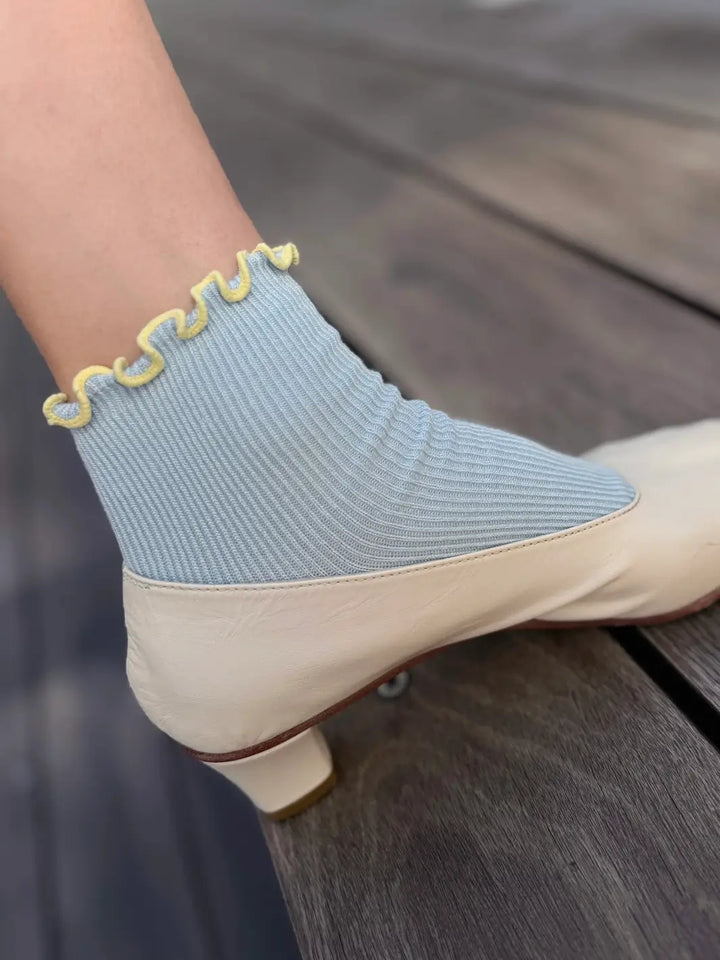 A close-up shot showing a foot wearing a cream-coloured kitten heel paired with The Petite Ruffled Modal Socks in Baby Blue / Butter from Le Bon Shoppe resting on a wooden deck | Twentyseven Toronto