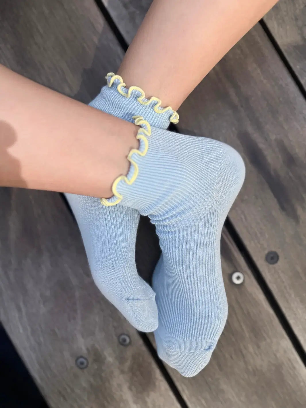 A top-down view captures a pair of feet crossed at the ankles, showcasing the baby blue ribbed texture and yellow contrast lettuce trim edges of The Petite Ruffled Modal Socks in Baby Blue / Butter from Le Bon Shoppe | Twentyseven Toronto