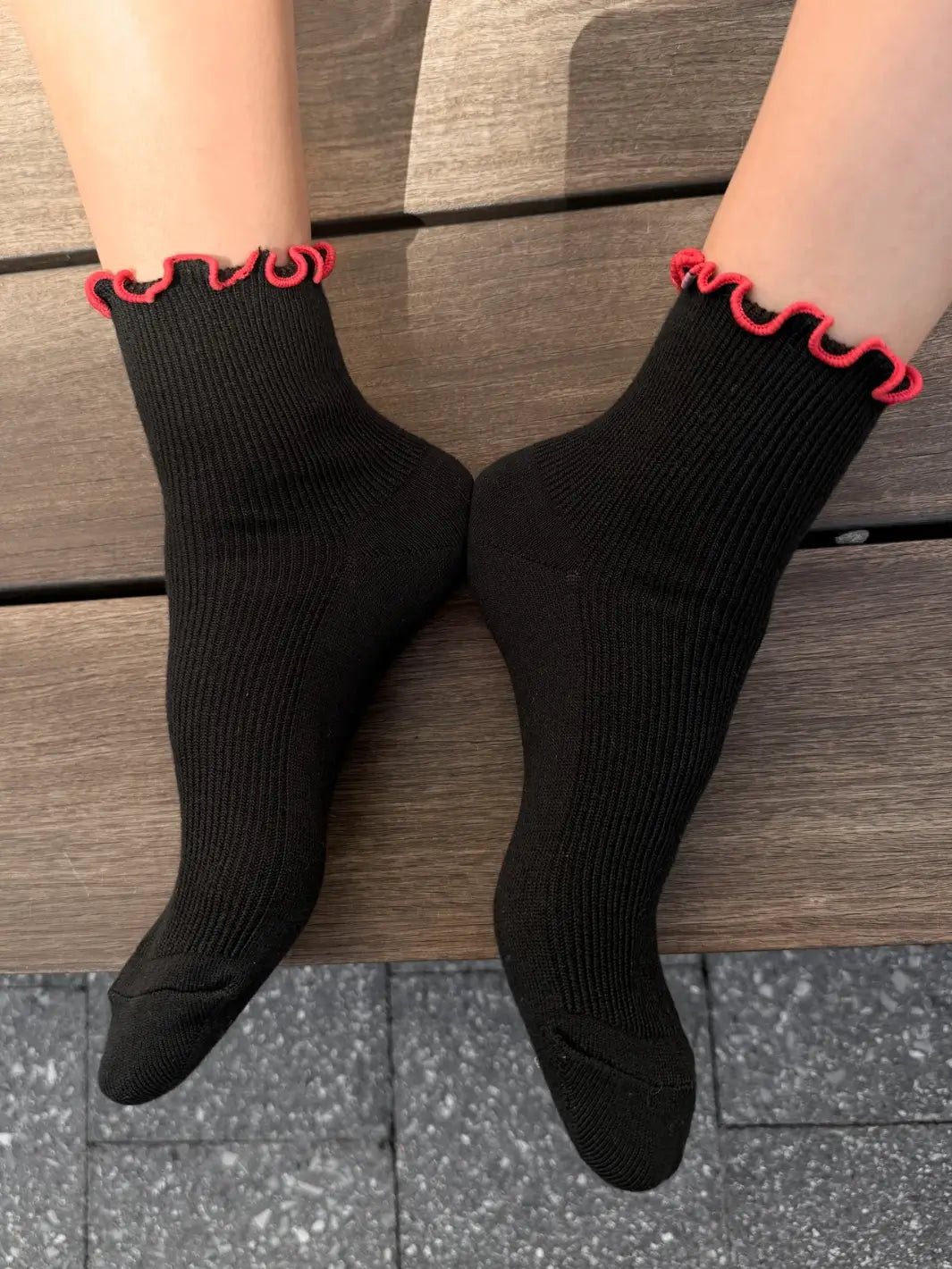 A close-up view from above shows a person's feet resting on a wooden bench while wearing The Petite Ruffled Modal Socks in Black / Red from Le Bon Shoppe | Twentyseven Toronto