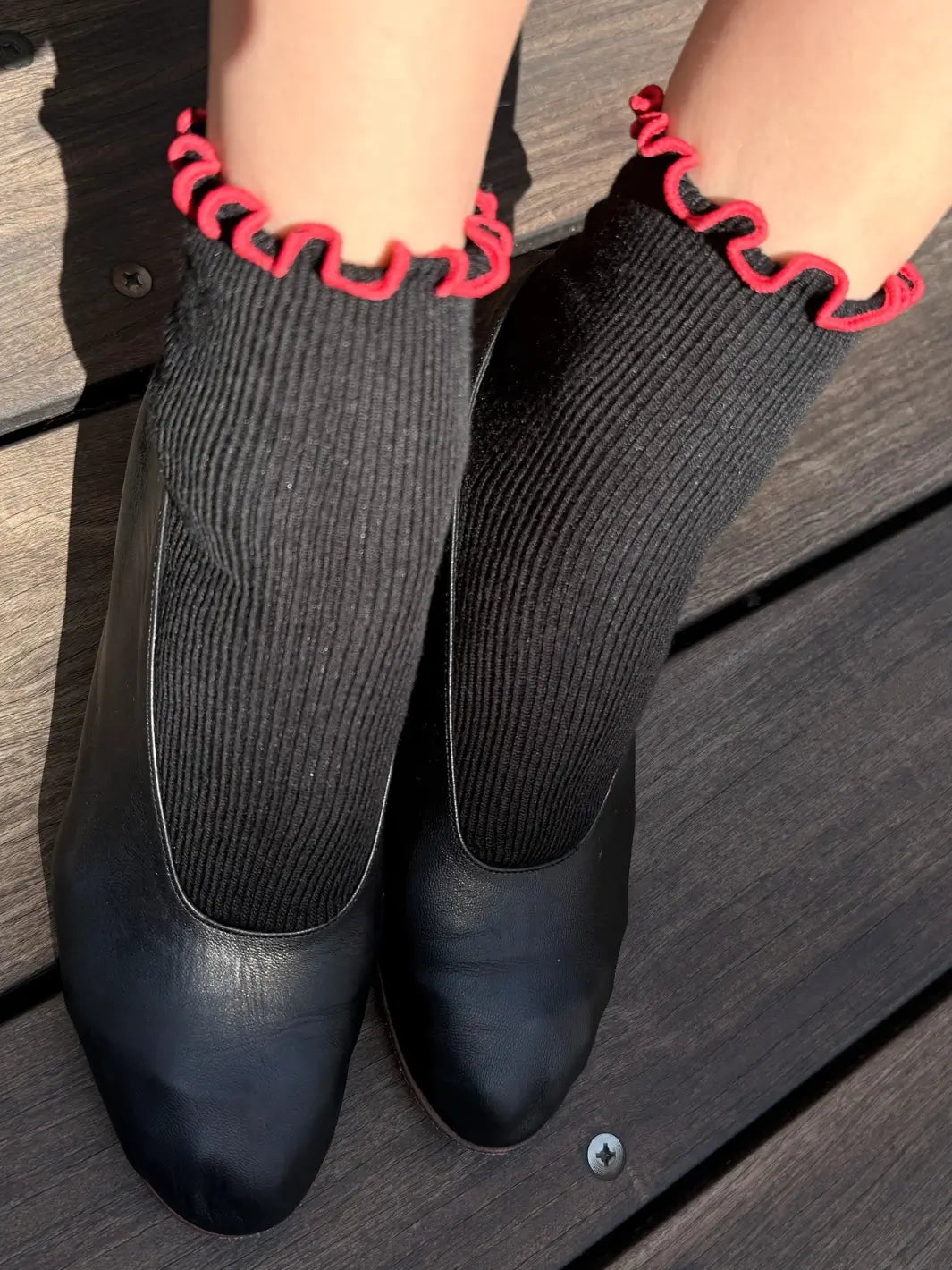 A person styles the distinct red lettuce-edge trim of The Petite Ruffled Modal Socks in Black / Red from Le Bon Shoppe with classic black leather ballet flats | Twentyseven Toronto