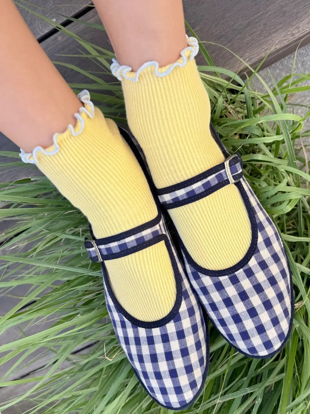 A high-angle shot showing The Petite Ruffled Modal Socks in Butter / Baby Blue from Le Bon Shoppe styled with blue-and-white checkered Mary Jane shoes in green grass | Twentyseven Toronto