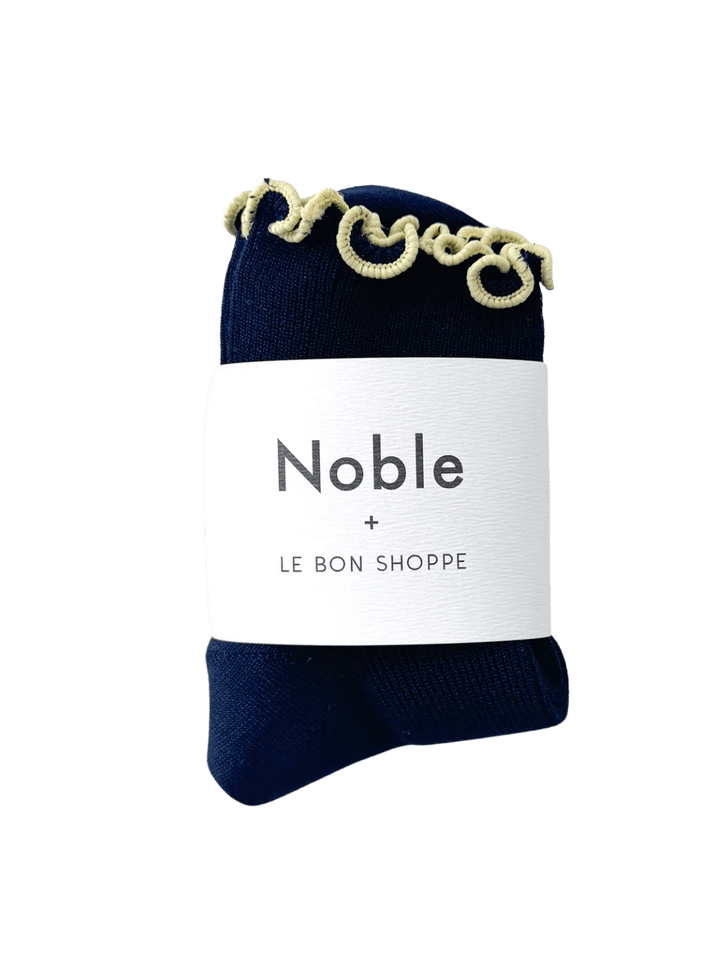 A navy sock with yellow edges of The Petite Ruffled Modal Socks in Butter / Navy from Le Bon Shoppe with a white paper brand band that reads "Noble + LE BON SHOPPE" | Twentyseven Toronto