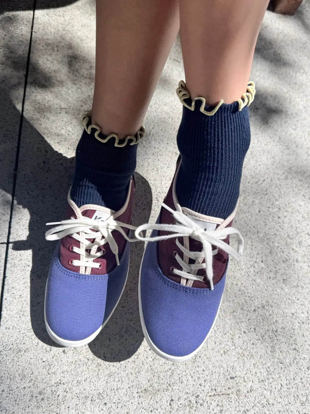 A person wears The Petite Ruffled Modal Socks in Butter / Navy from Le Bon Shoppe paired with blue and burgundy canvas sneakers on a concrete surface | Twentyseven Toronto
